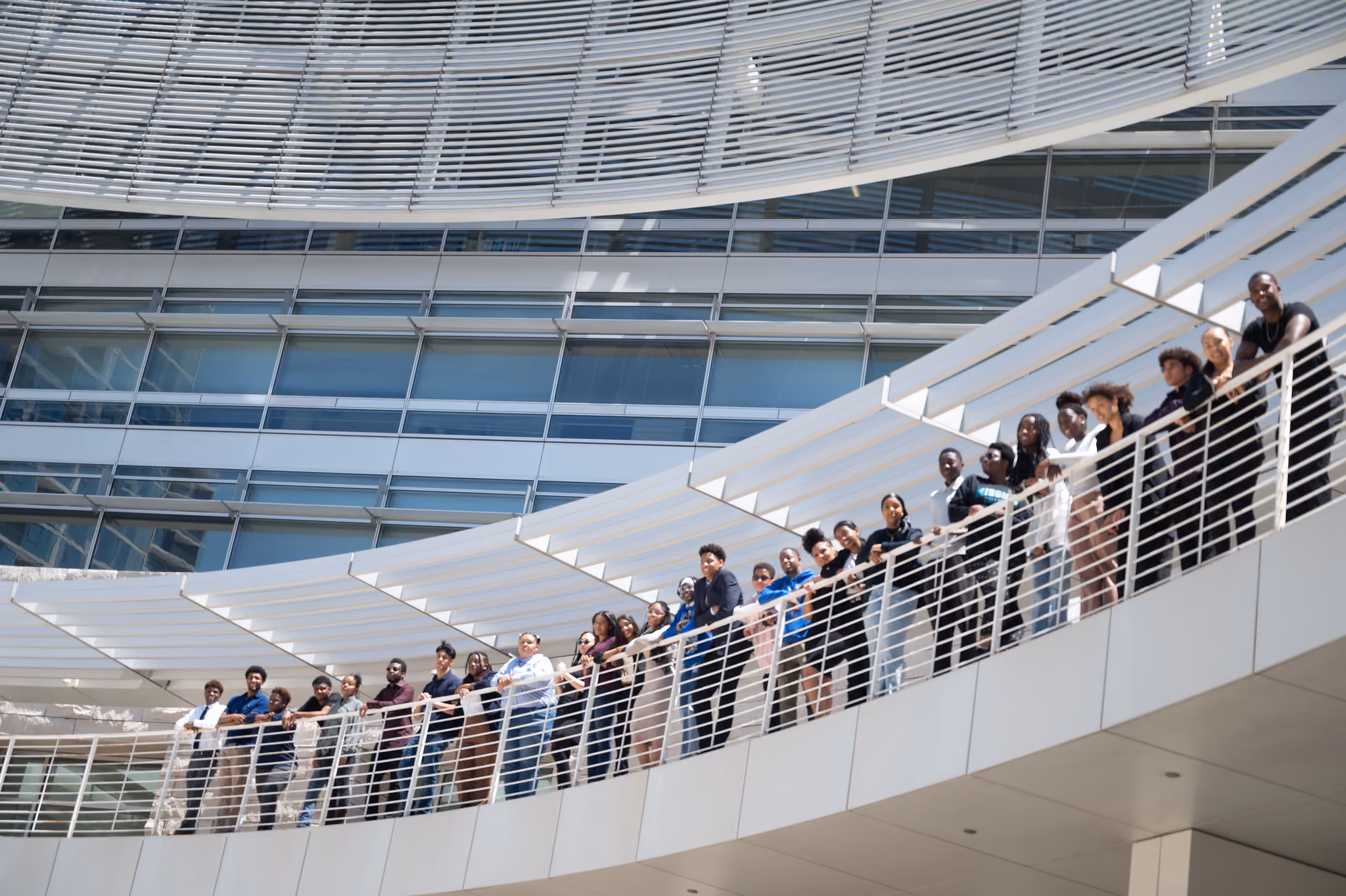 Participants line a curved white walkway outside a glass and stone building.