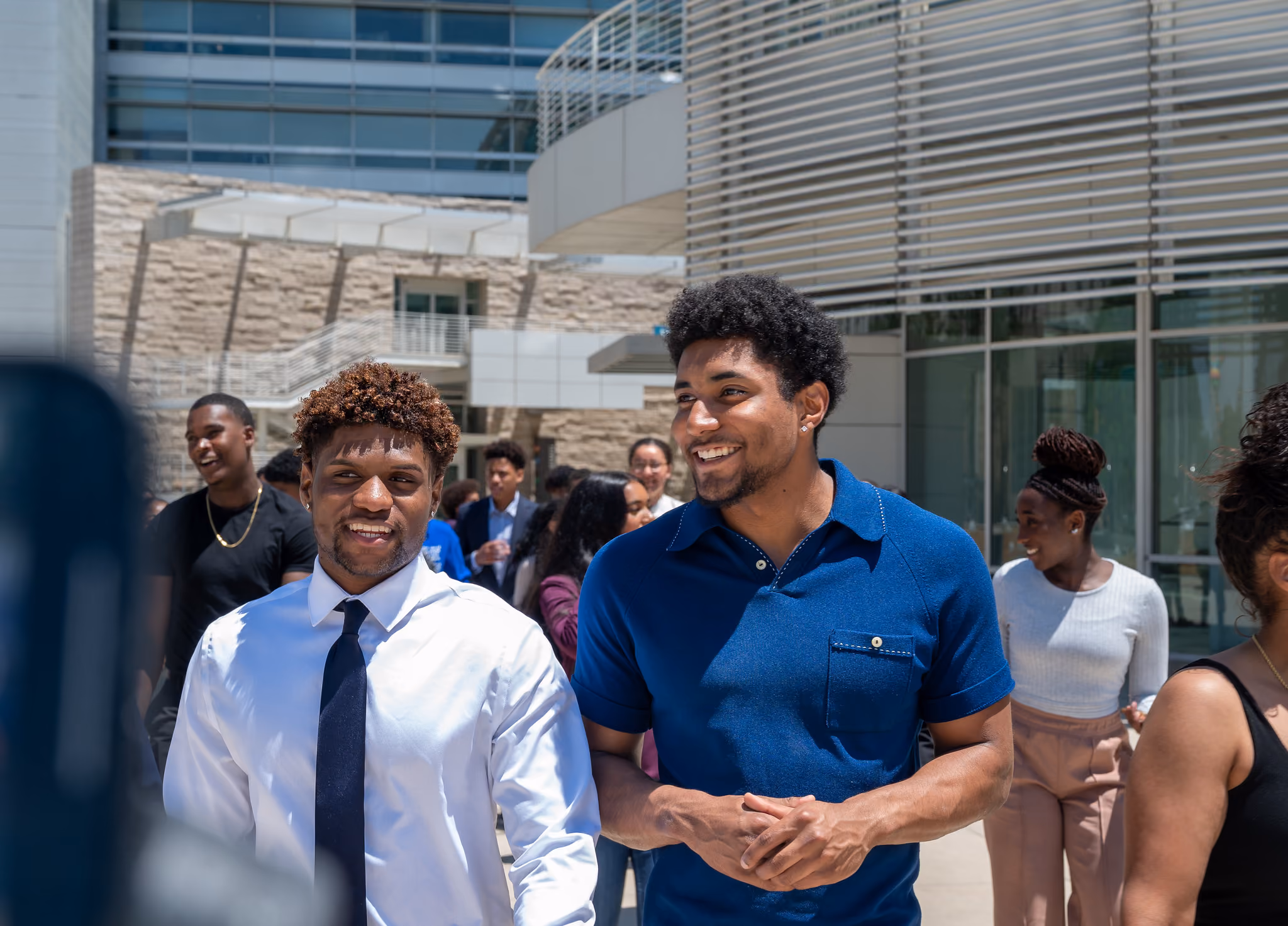 Two smiling young men, one in a shirt and tie and one in a blue polo, stand outdoors among a crowd.