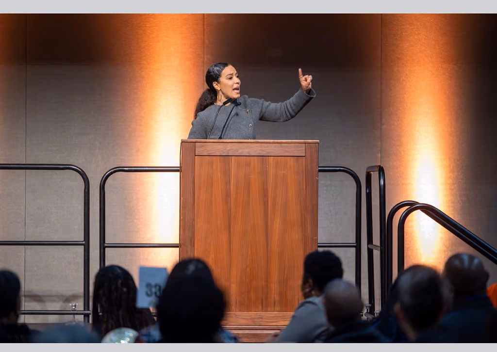 A speaker in a gray jacket raises one finger while addressing the audience from a wooden podium