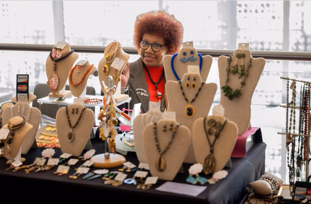 A smiling vendor with glasses sits behind a table filled with necklaces, earrings, and jewelry displays
