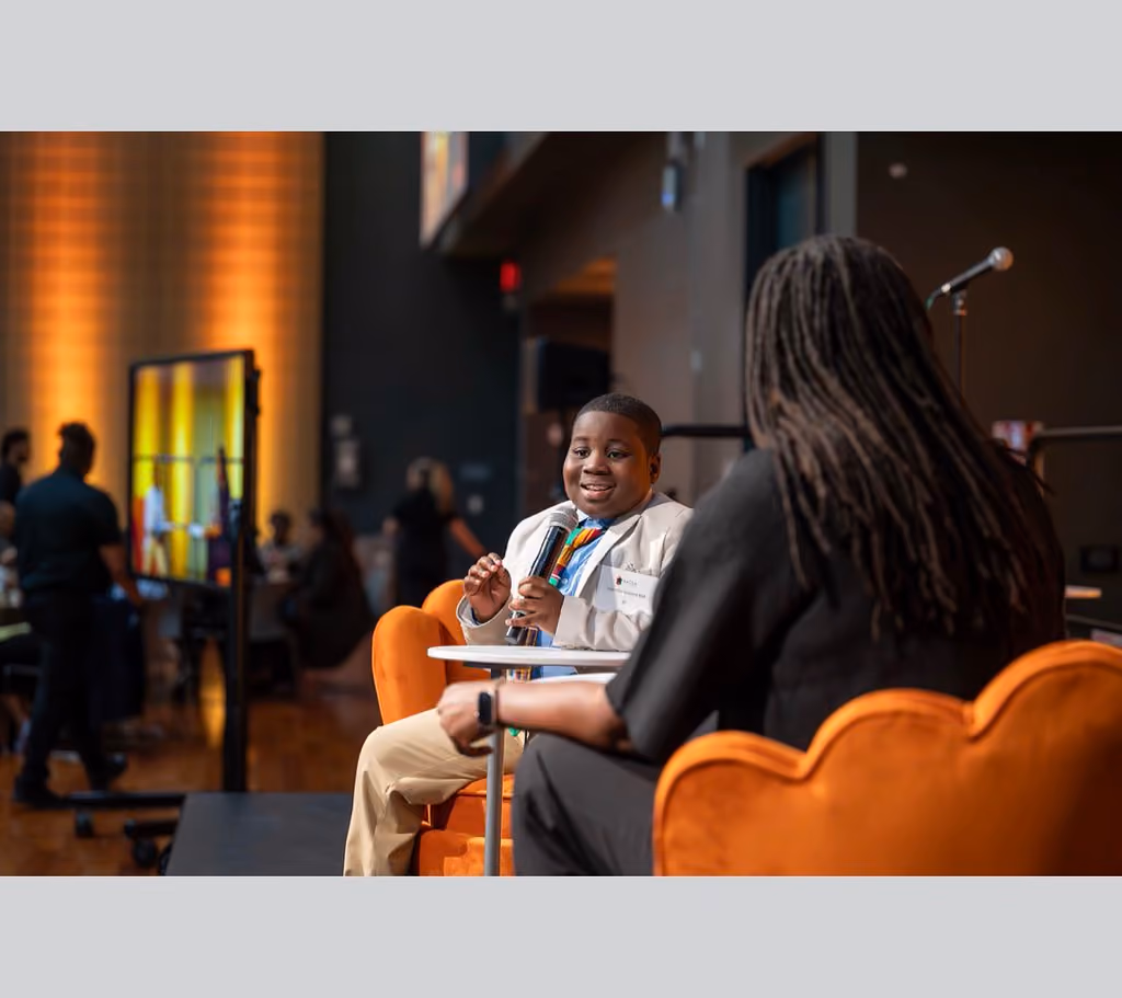 A speaker in a light blazer holds a microphone while facing another speaker seated in an orange armchair