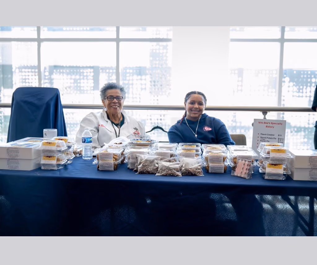 Two smiling vendors sit behind a table covered with packaged baked goods and a bakery menu sign