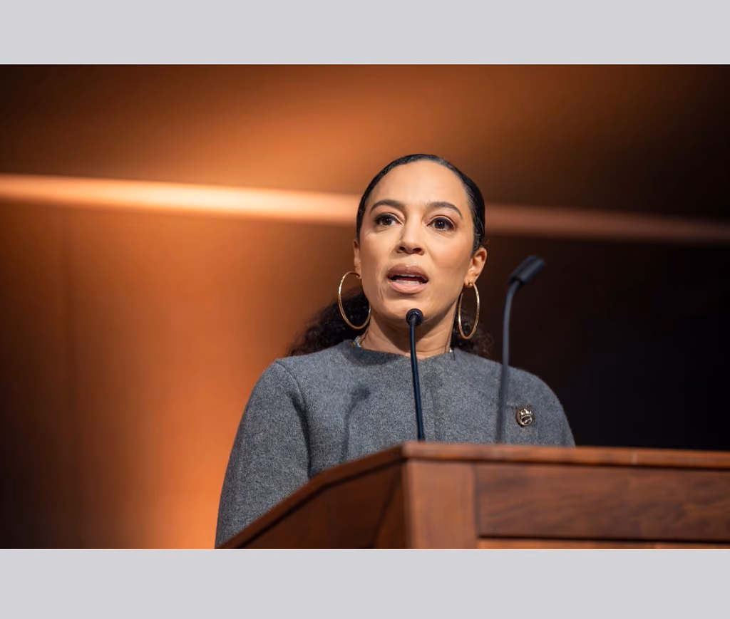 A speaker wearing hoop earrings and a gray jacket addresses the room from behind a wooden podium