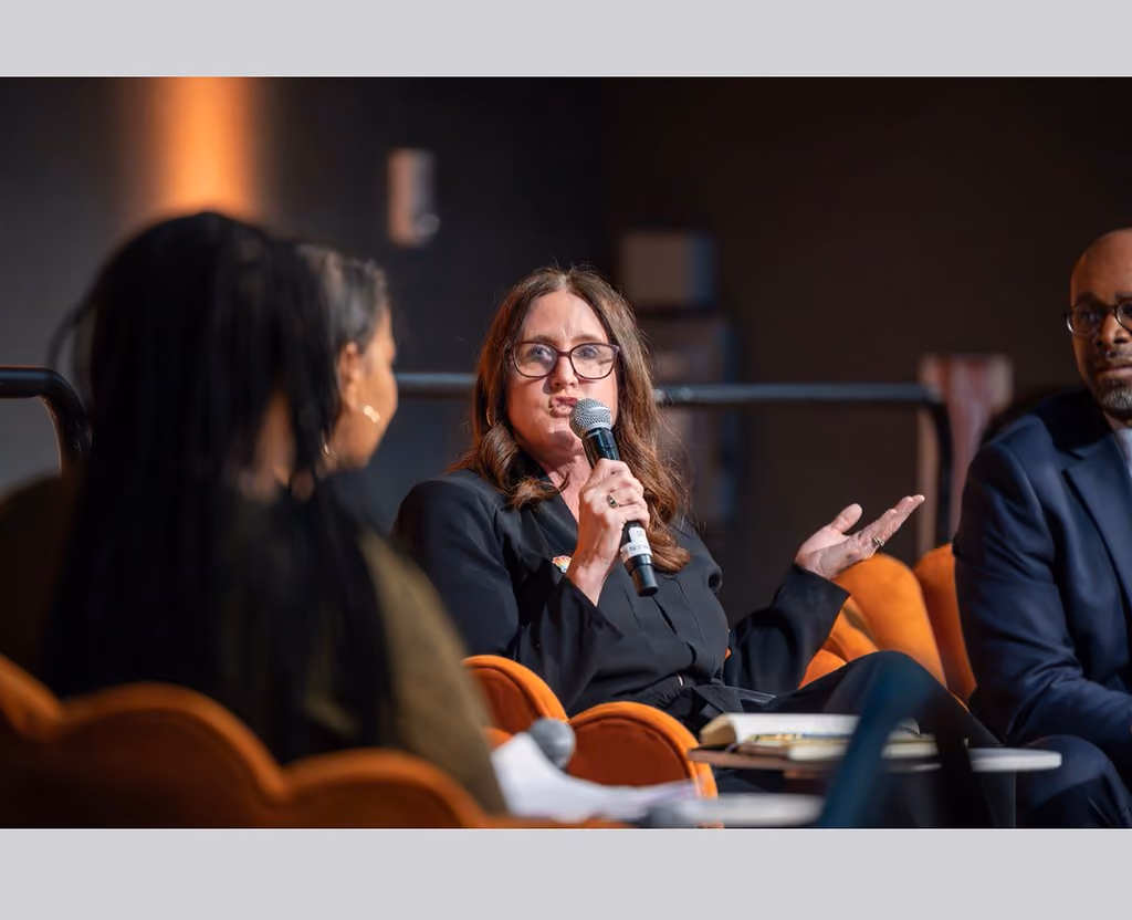 A seated panelist in black speaks into a handheld microphone and gestures with an open palm