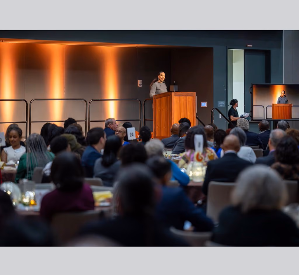 Guests seated at banquet tables face a speaker at a wooden podium, with a live video display beside the stage