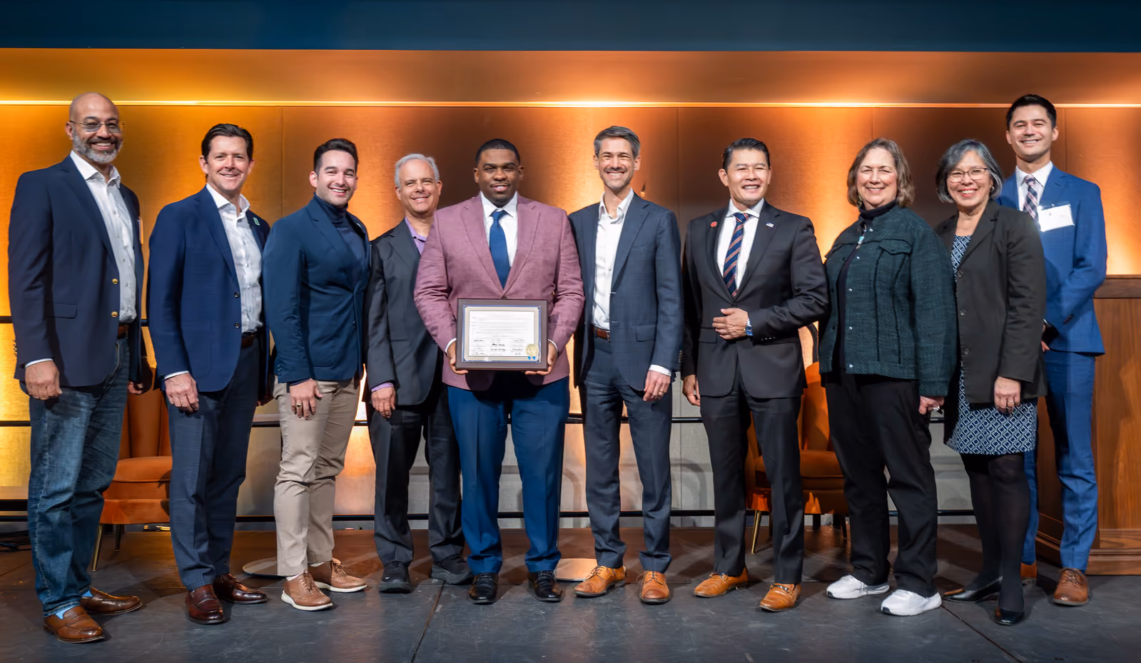 Ten guests pose onstage as a guest in a pink blazer holds a framed certificate against an amber-lit wall