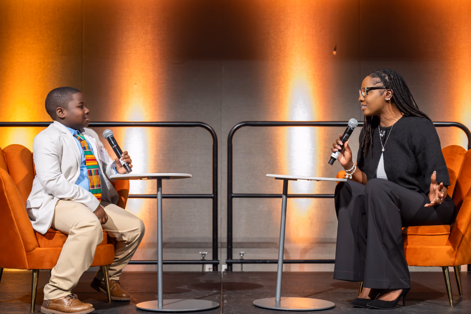 Two speakers face each other with handheld microphones while seated in orange armchairs onstage