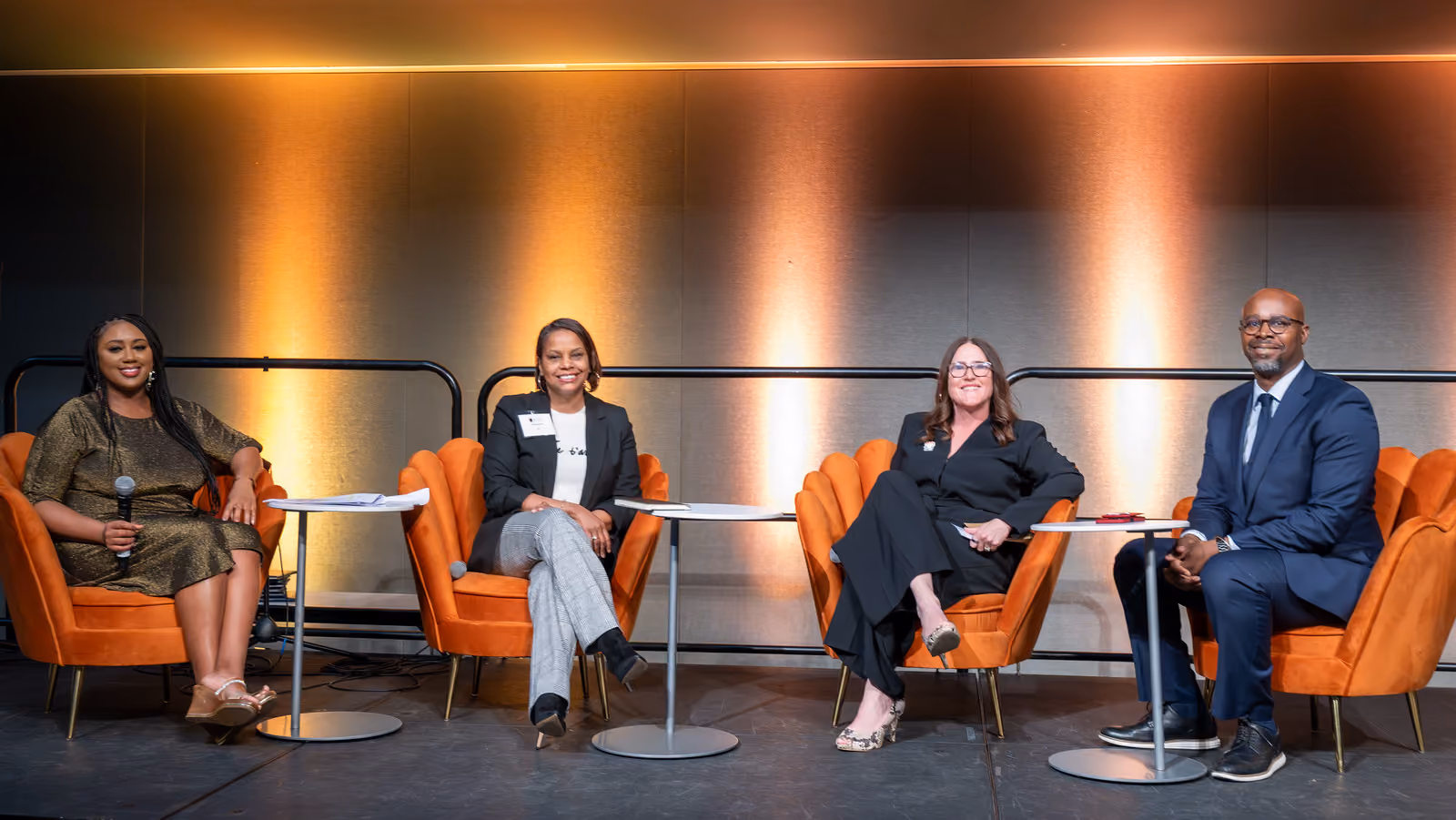 Four panelists smile from orange armchairs arranged with small round tables on an amber-lit stage