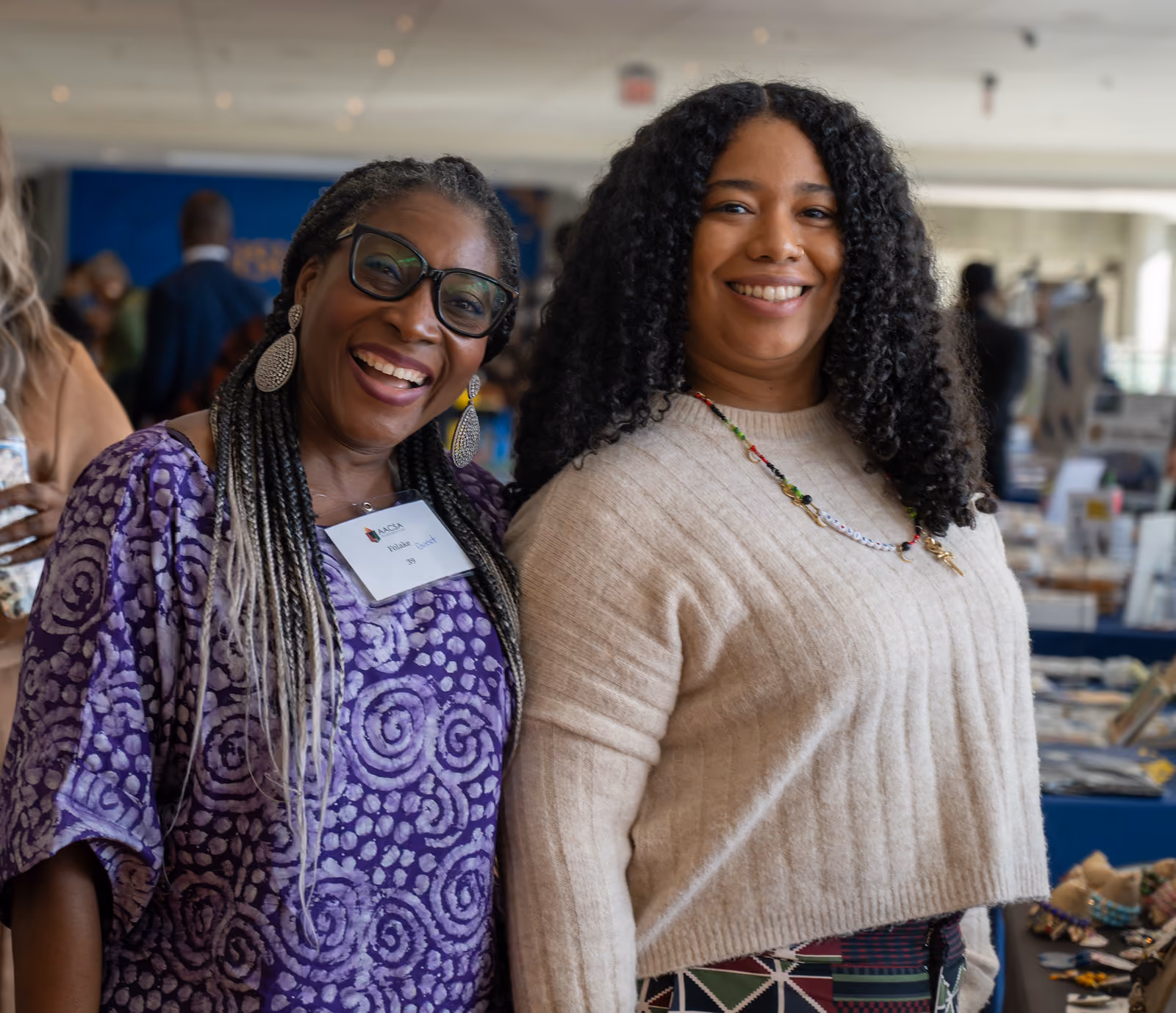 Two smiling guests, one in a purple patterned outfit and one in a cream sweater, pose beside vendor tables