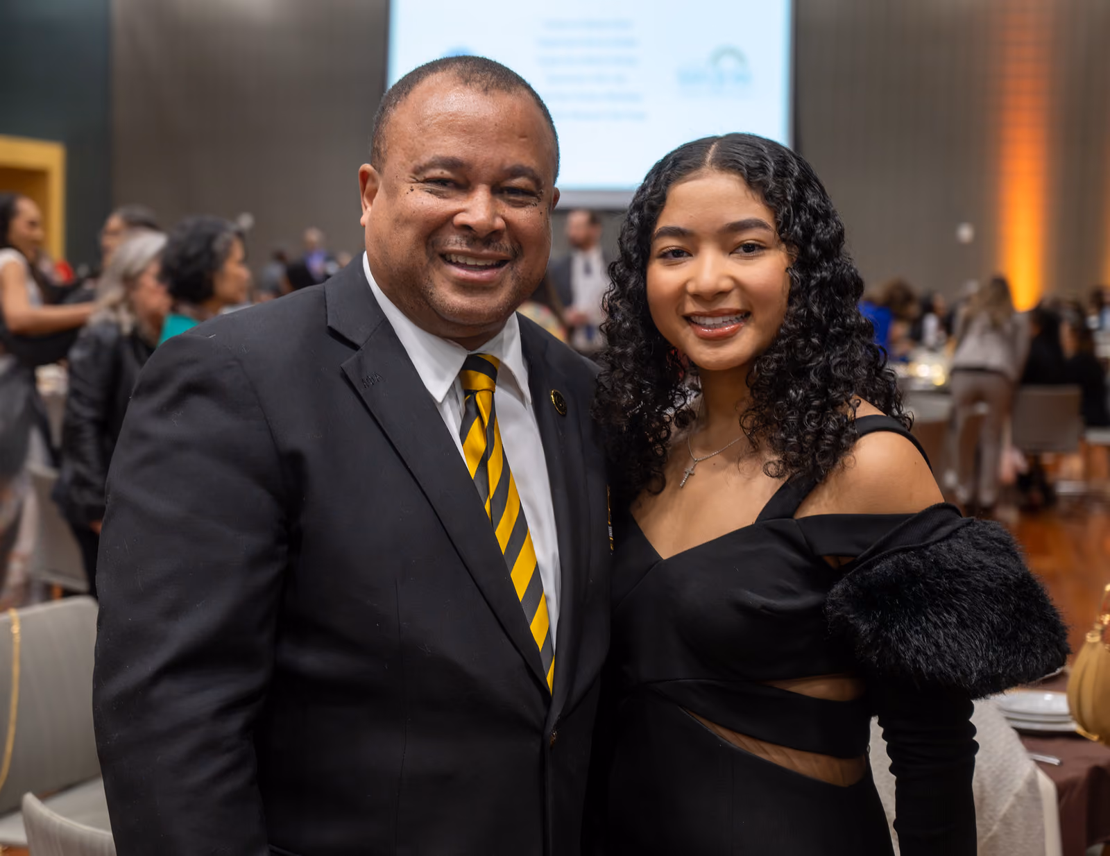 Two smiling guests pose together in formal black attire beside banquet tables in the luncheon hall