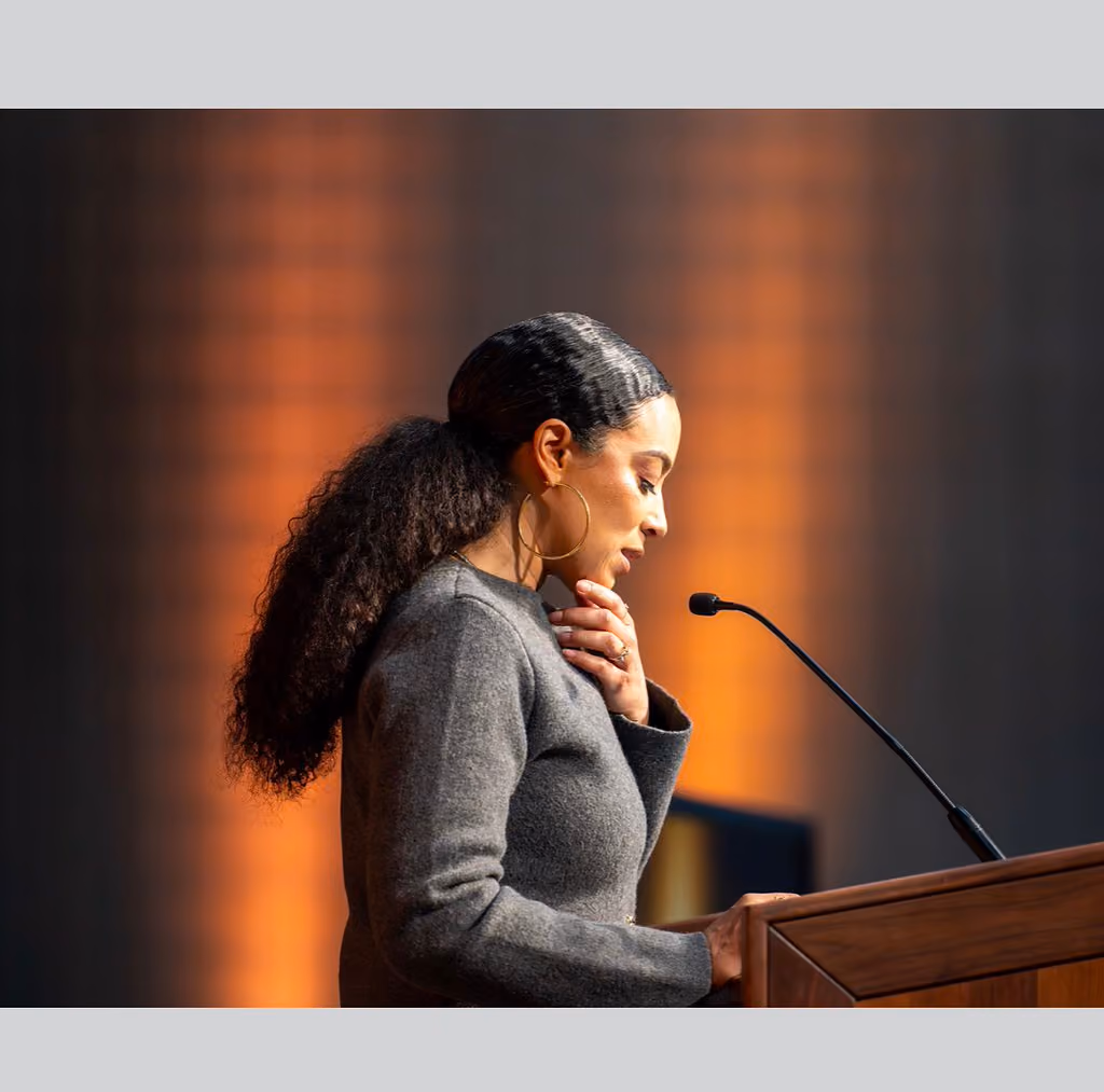 A speaker in a gray jacket looks down at the wooden podium with one hand resting near their collar