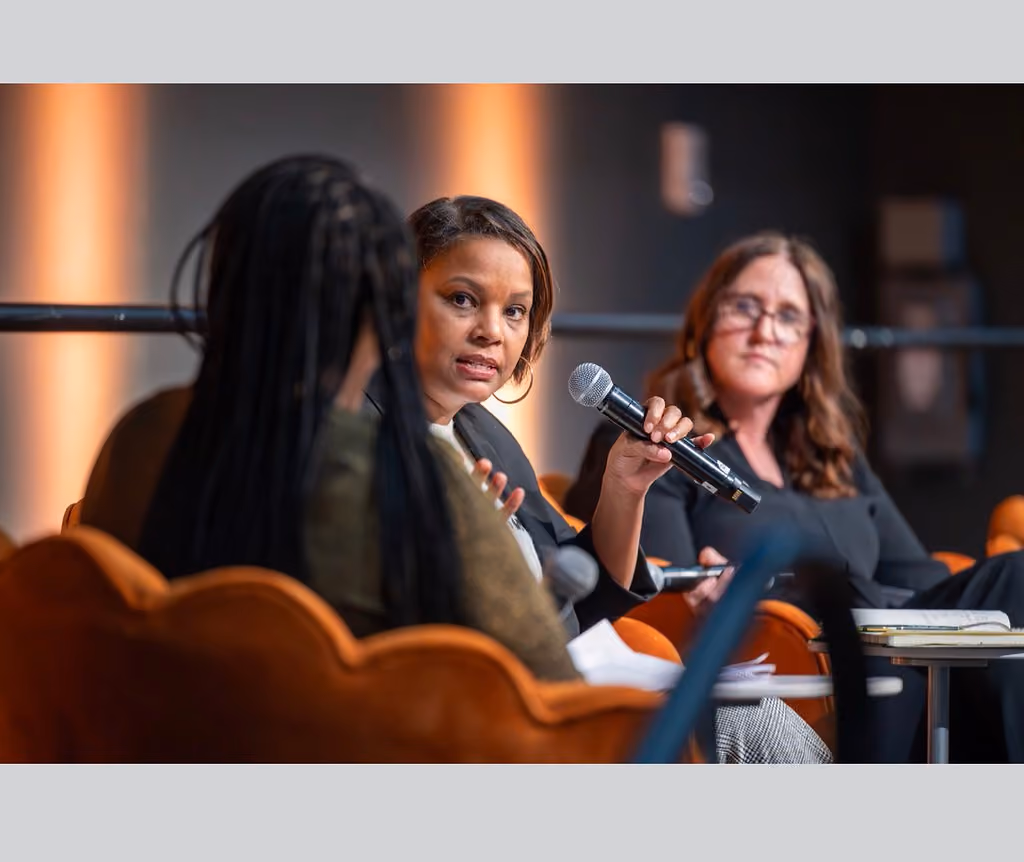 A panelist in a black blazer holds a microphone and addresses another panelist seated in the foreground