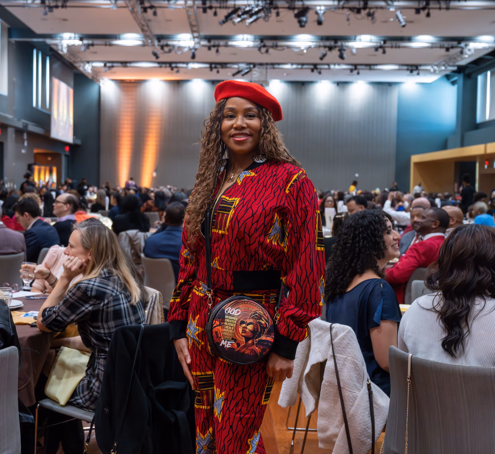 A guest in a red beret and matching patterned outfit poses among seated guests in the luncheon hall