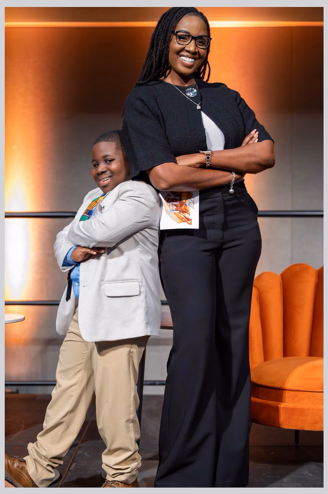 Two smiling participants pose back to back with folded arms beside an orange armchair onstage