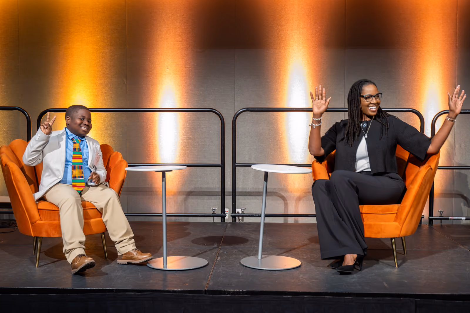 Two seated speakers smile and raise their hands from orange armchairs on an amber-lit stage