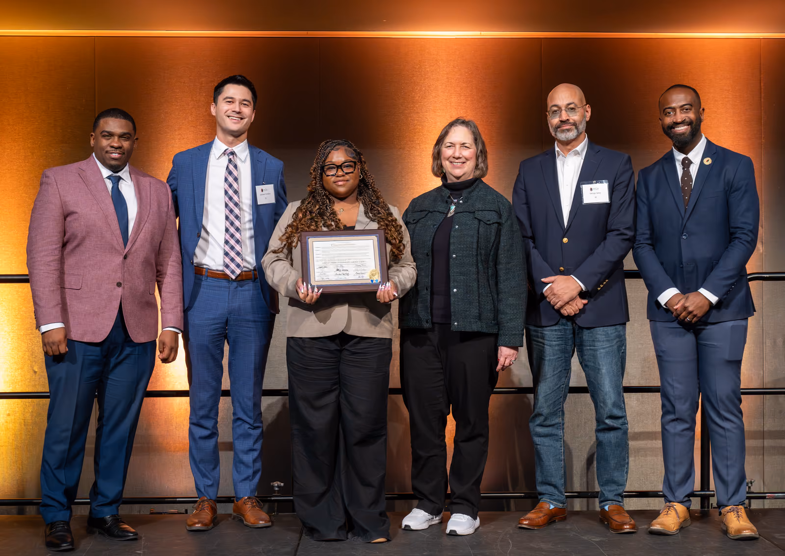 Six guests stand together on an amber-lit stage as the central guest displays a framed certificate
