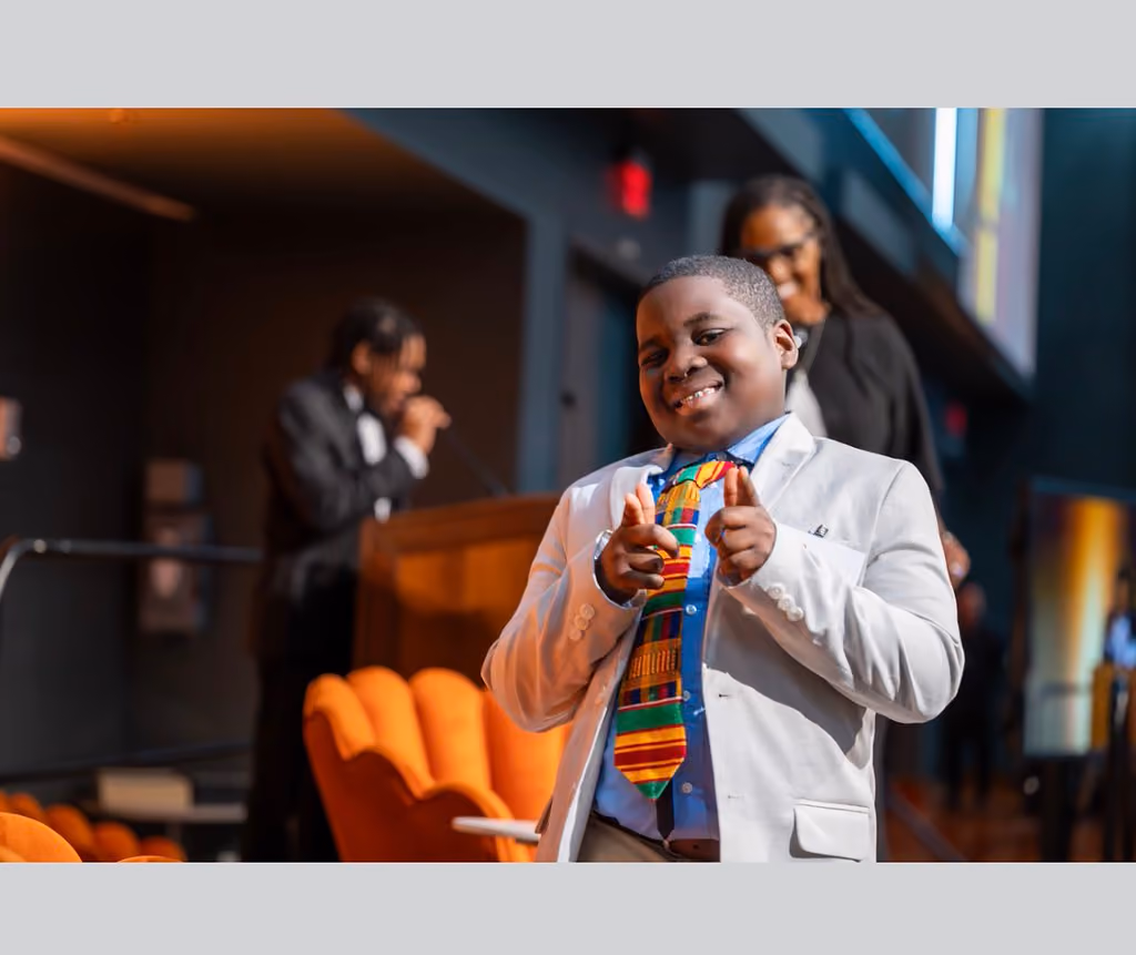 A smiling participant in a light blazer and colorful tie points both hands toward the camera near the stage