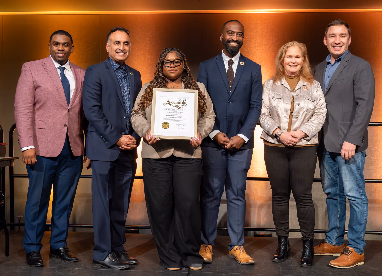 Six guests pose onstage with a guest holding a large framed certificate at the center