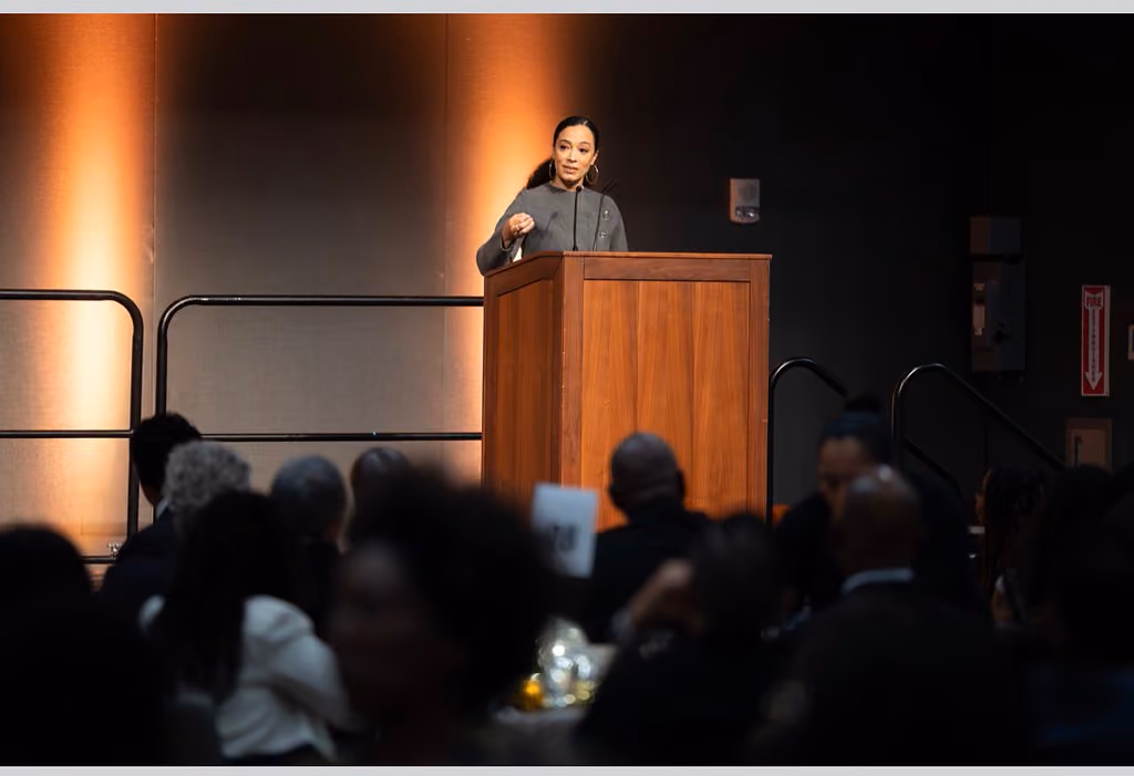 A speaker in a gray jacket gestures from a wooden podium as guests sit at banquet tables in the foreground
