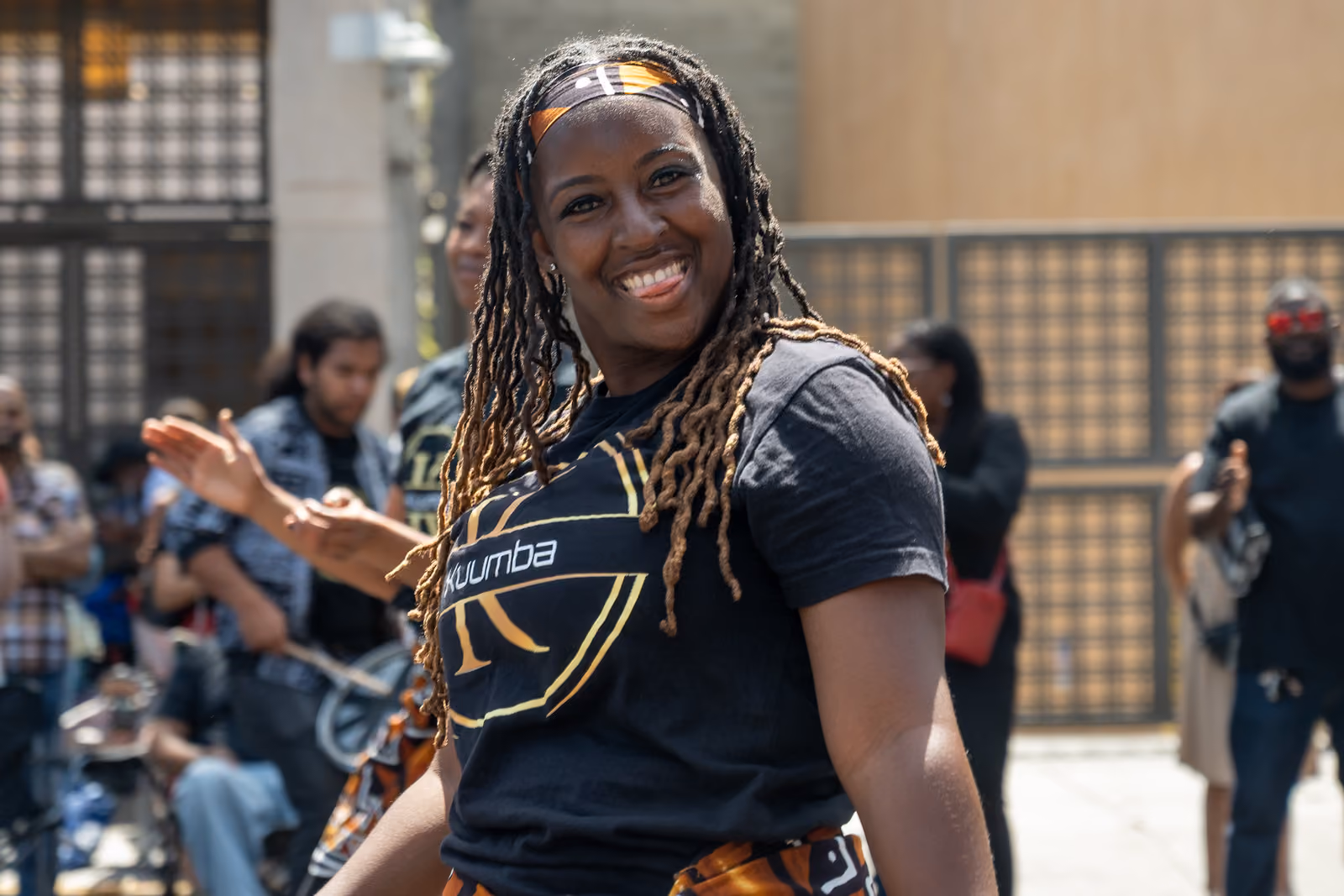 A smiling dancer with long locs and a patterned headband poses among festival attendees
