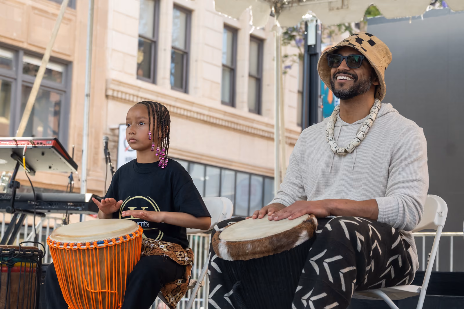 Two seated drummers play hand drums on an outdoor stage beside keyboards and microphones