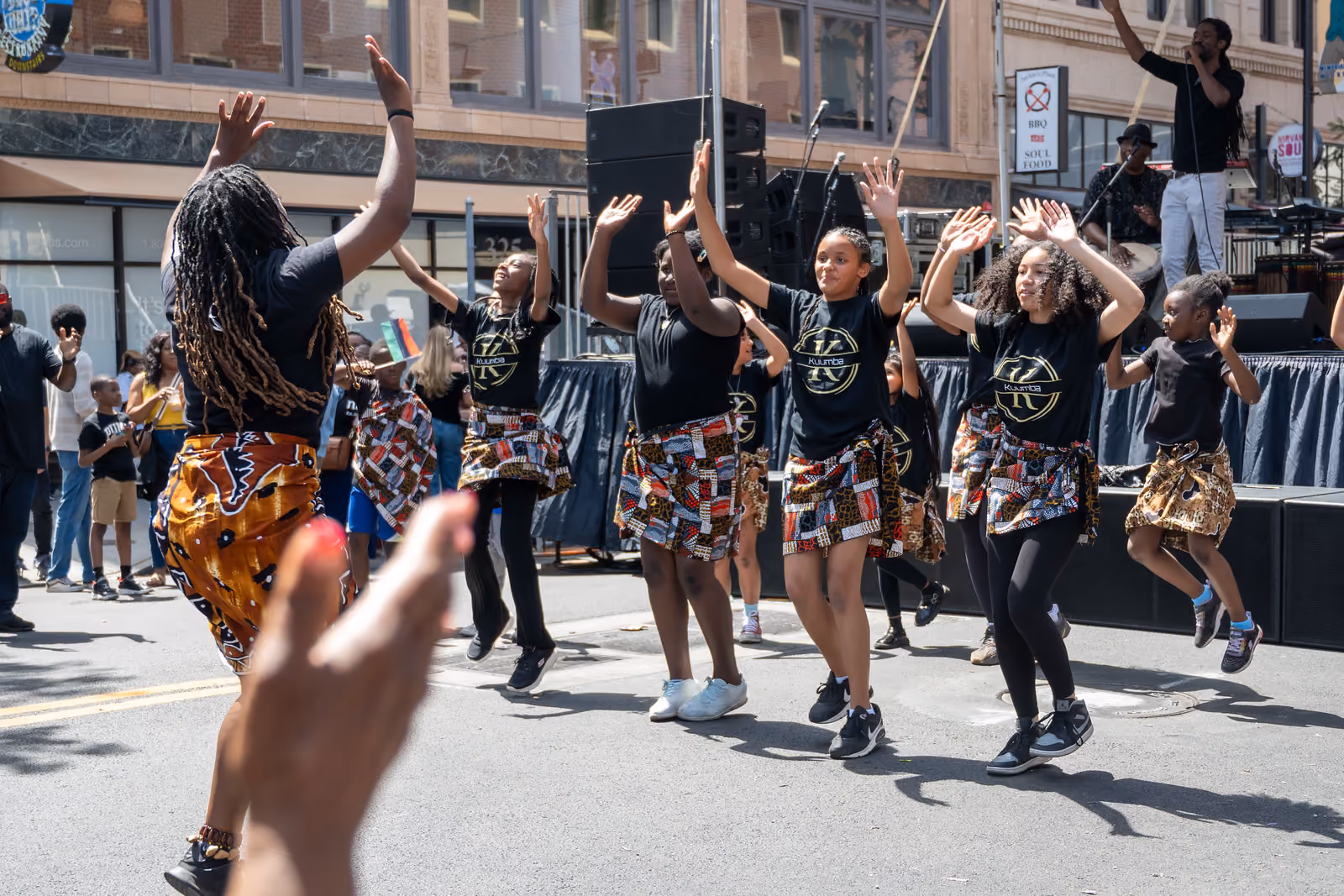 A dance group in black shirts and patterned wraps raises their hands beside an outdoor stage