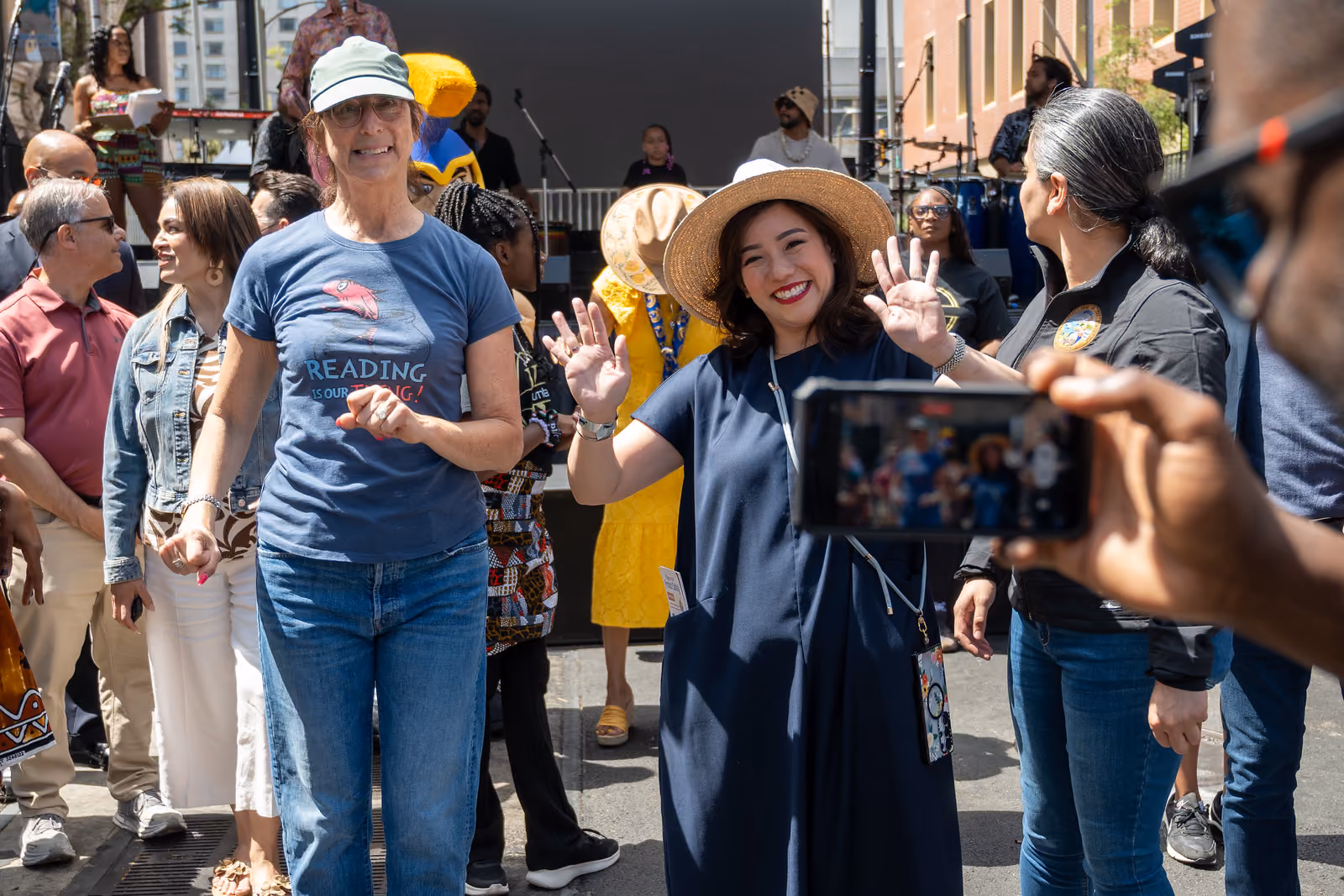 Festival guests gather near the stage as a woman in a wide-brimmed hat waves toward a camera