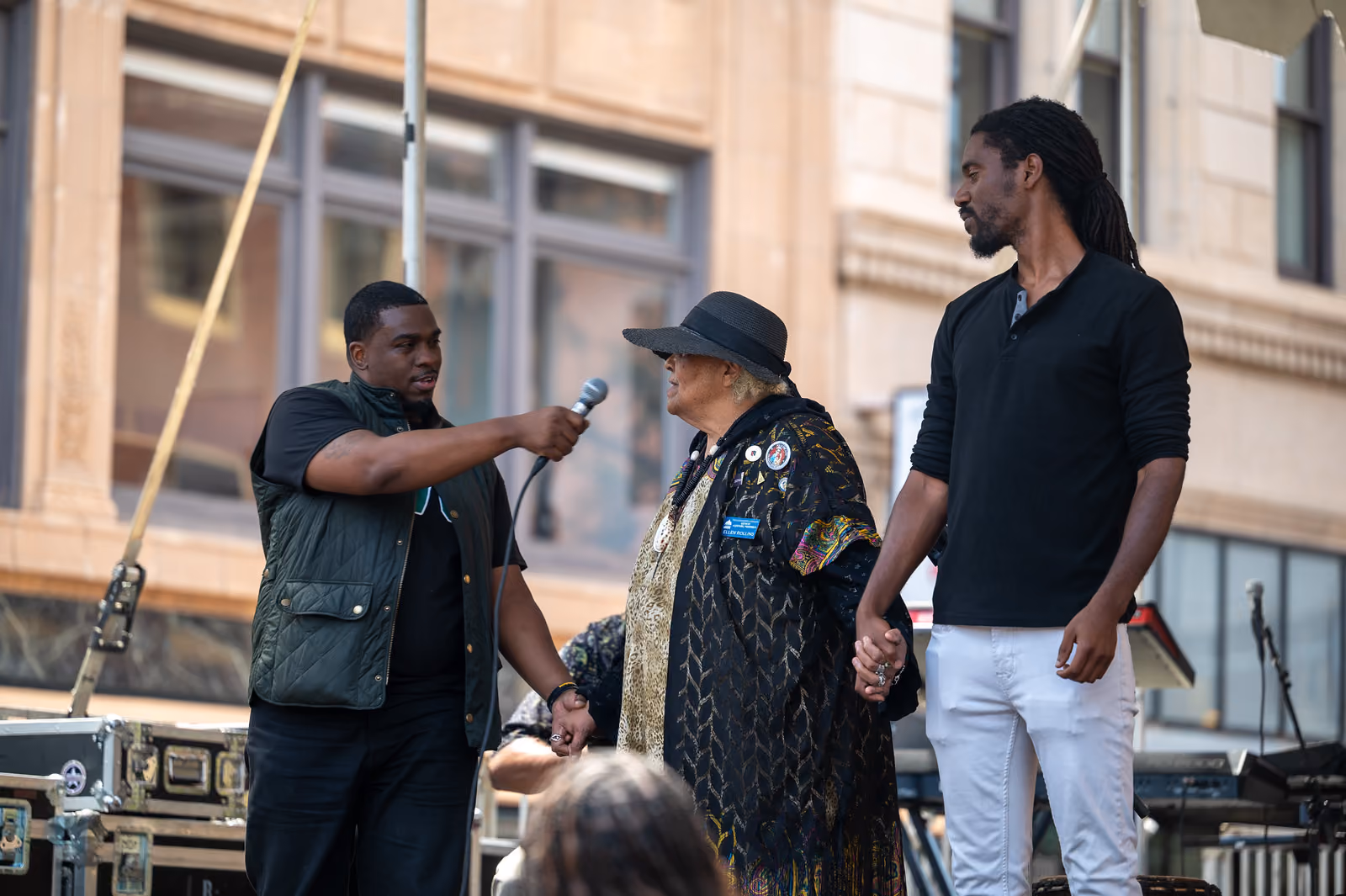 A microphone holder interviews a guest in a black hat who holds hands with a man on the outdoor stage