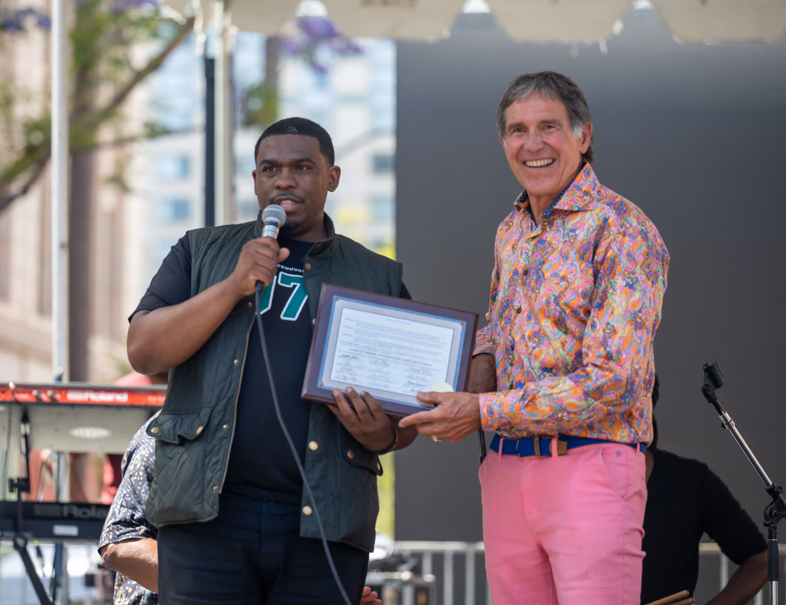 Two men hold a framed certificate on an outdoor stage while one speaks into a microphone
