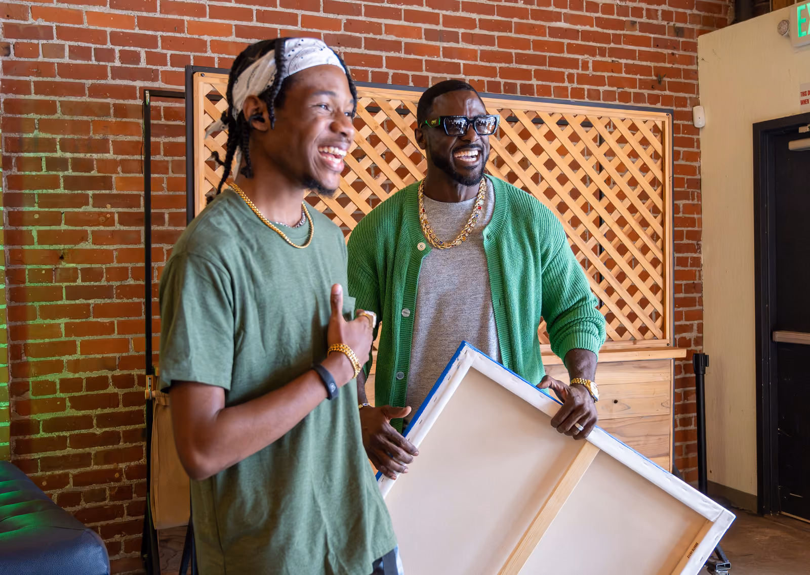 Two smiling men pose indoors against a brick wall, one holding a large canvas with its back facing out