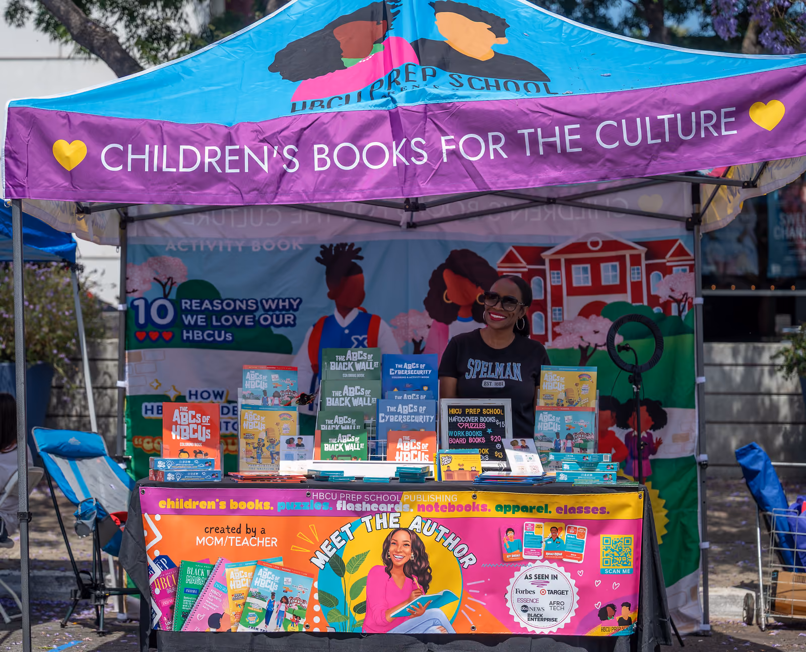 A bookseller stands behind colorful children's books beneath a tent reading Children's Books for the Culture