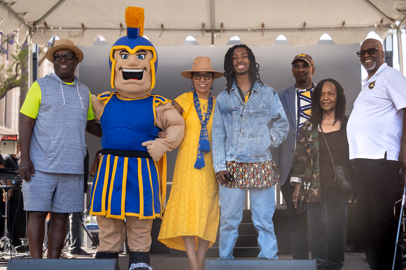 Six guests and a blue-and-gold Spartan mascot pose together beneath the outdoor stage canopy