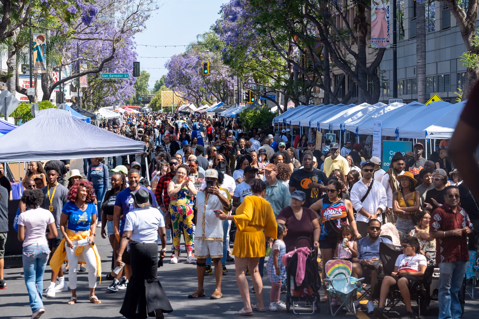 A large festival crowd fills a street lined with vendor tents and purple flowering trees