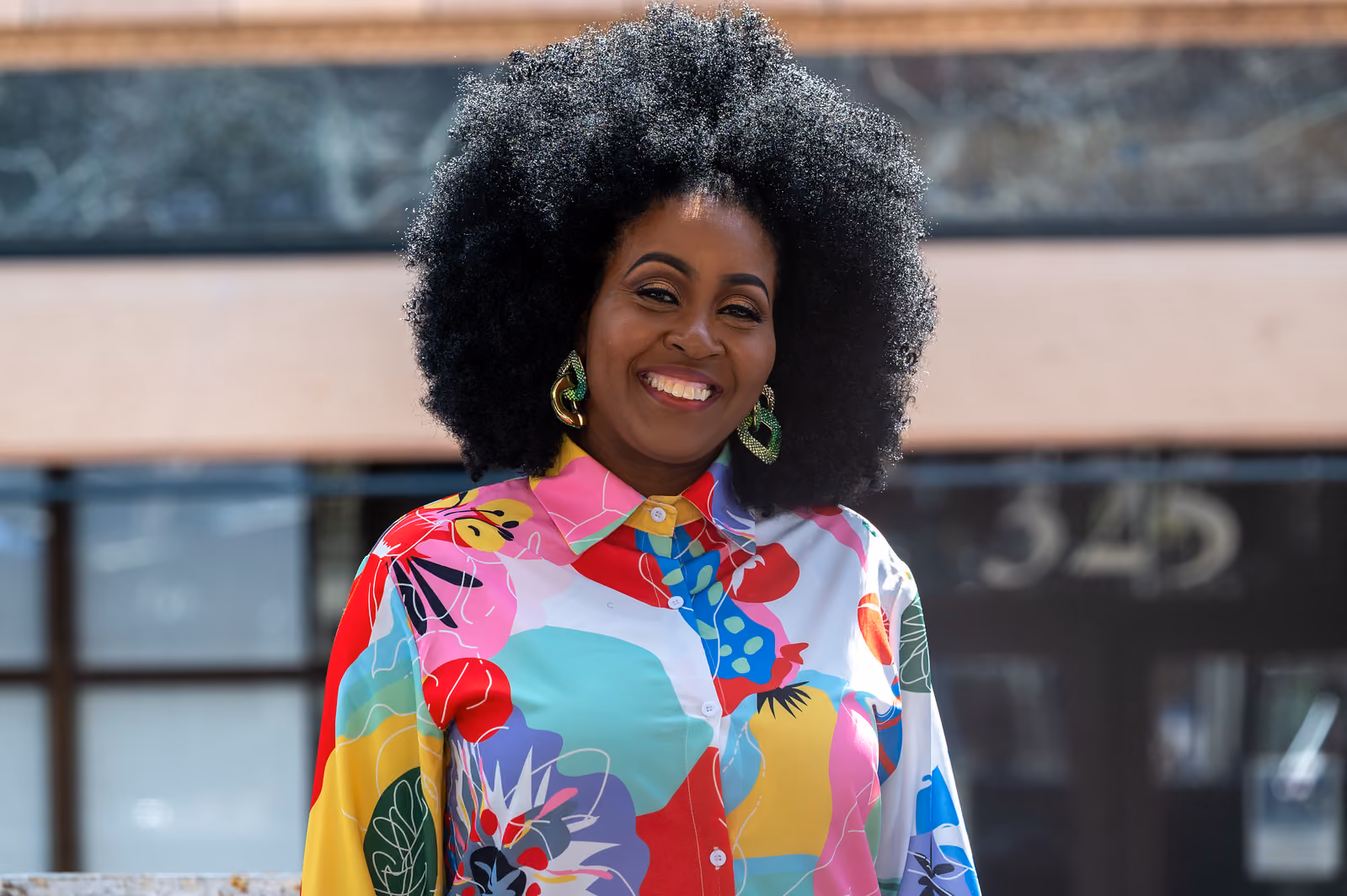 A smiling woman with a full afro and colorful floral shirt poses outdoors in front of a building