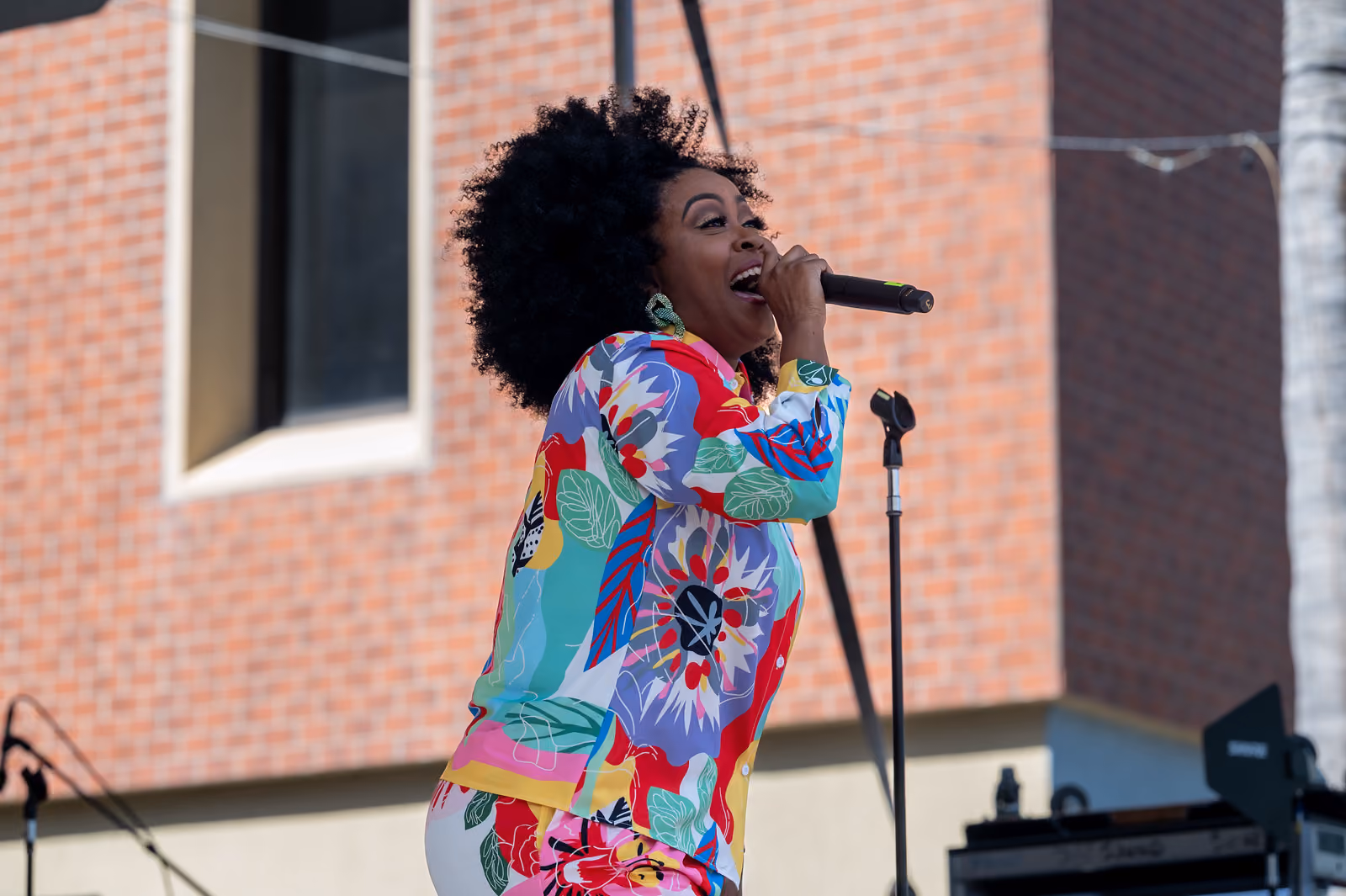A singer in a colorful floral outfit performs with a handheld microphone on an outdoor stage