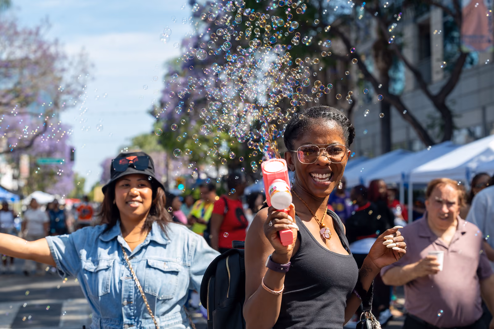 A smiling festival attendee holds a pink bubble gun as bubbles float above the crowded street