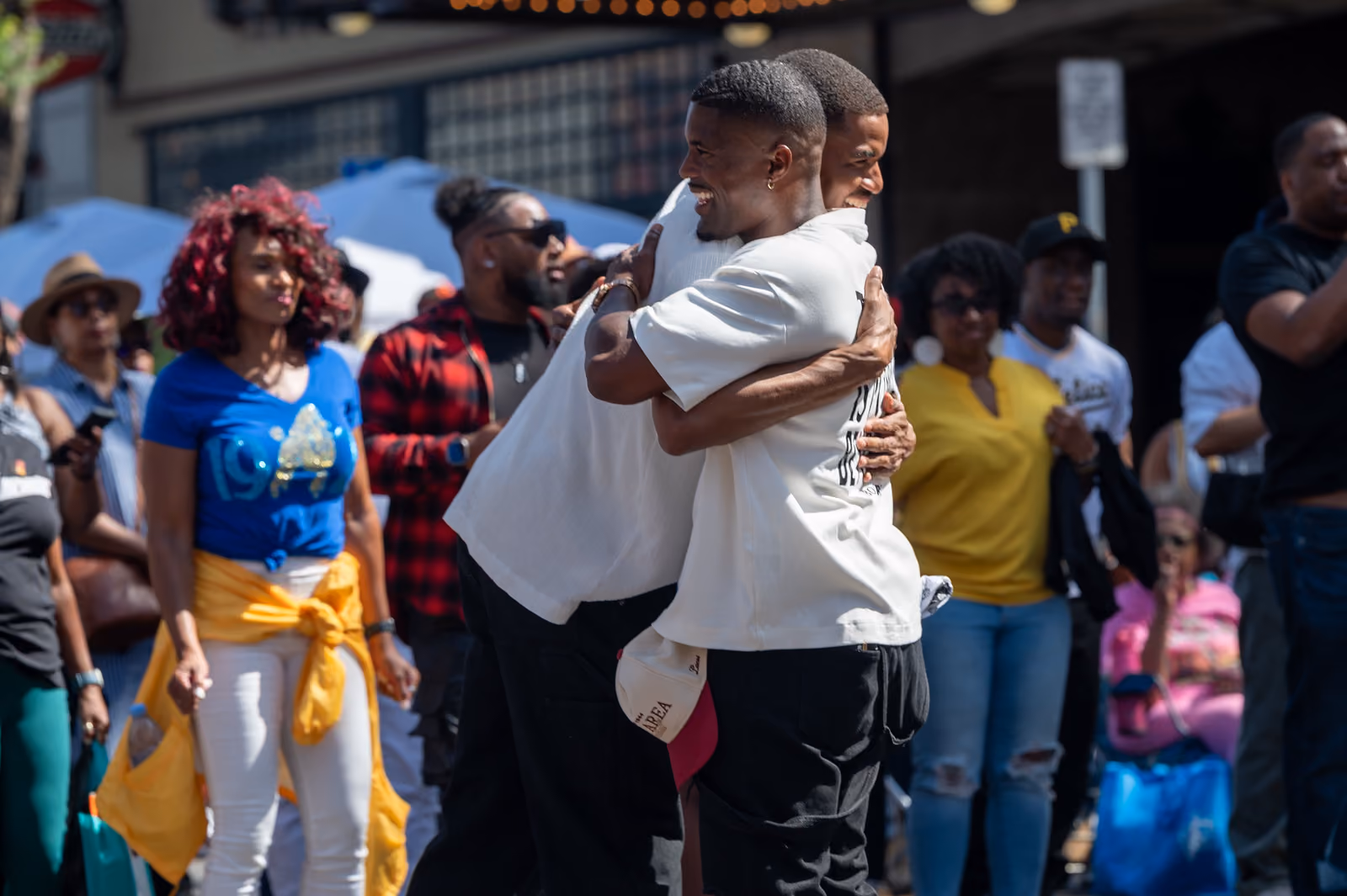 Two smiling festival attendees embrace in the street as a crowd watches behind them