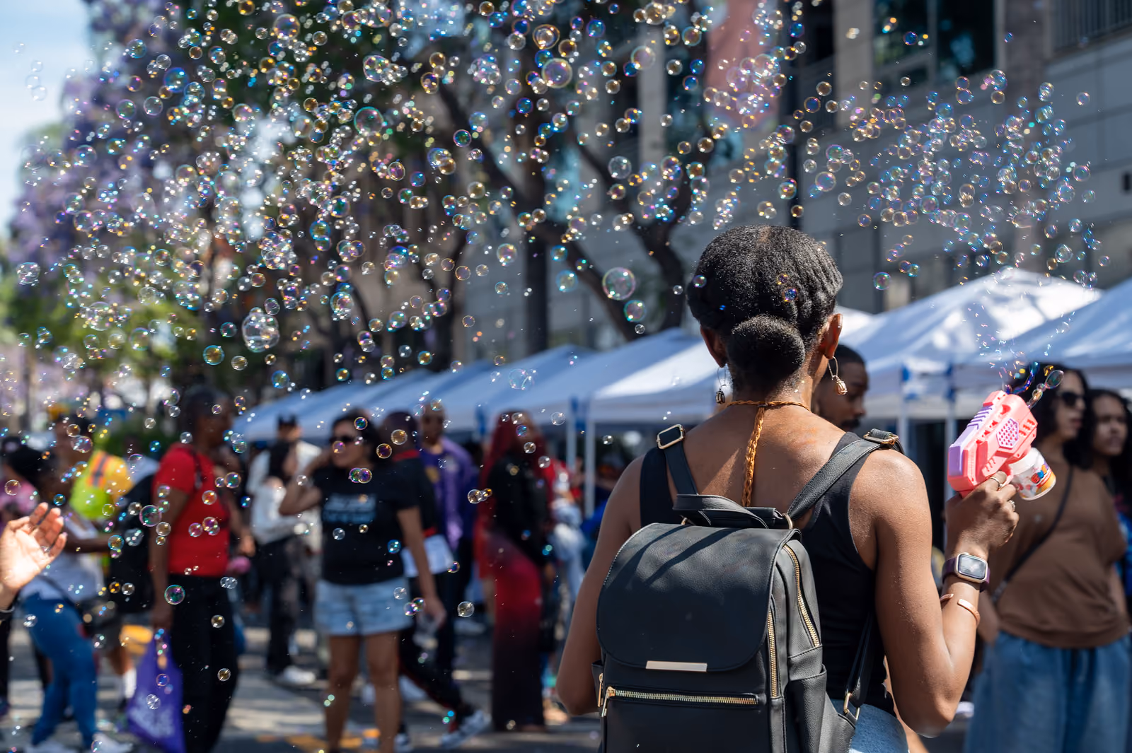 An attendee with a black backpack holds a pink bubble gun beneath bubbles beside vendor tents