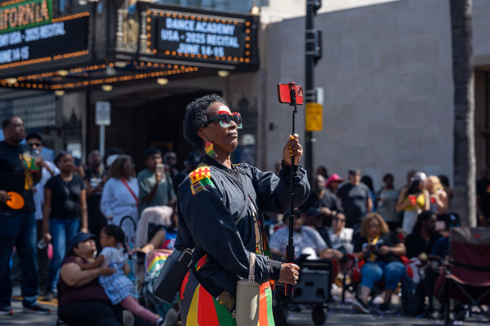 An attendee wearing colorful sunglasses records with a phone on a selfie stick near the theater