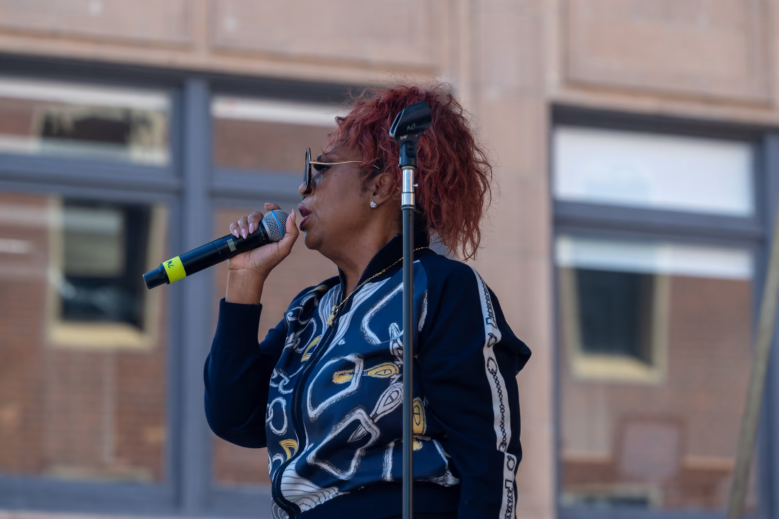 A singer with reddish hair and sunglasses holds a microphone beside a stand on an outdoor stage