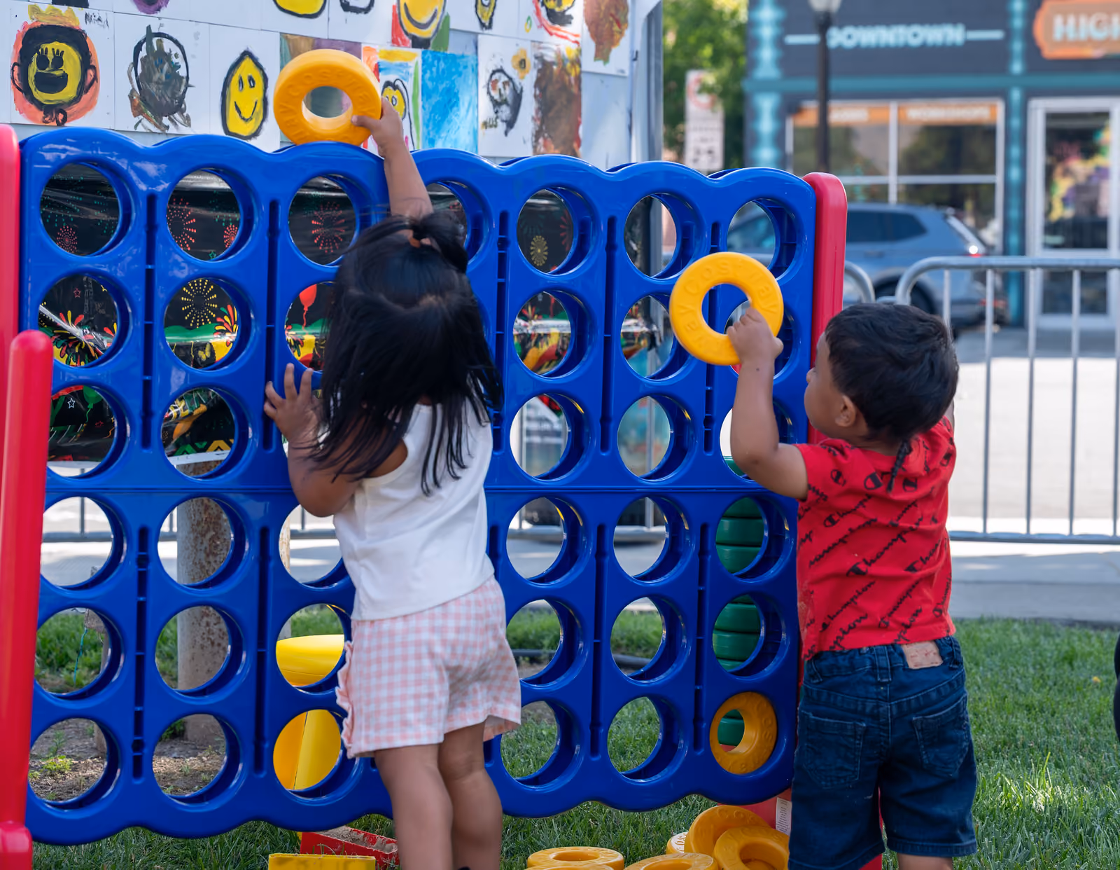 Two children reach up to place yellow rings into a giant blue four-in-a-row game outdoors