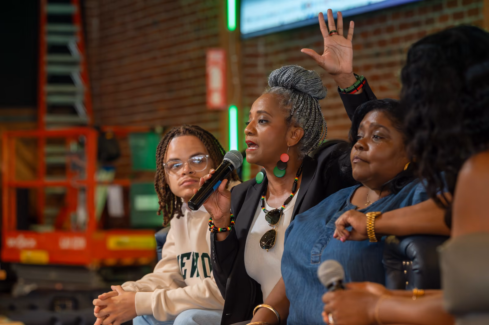 A seated panelist with gray braids speaks into a microphone and raises a hand beside other panelists