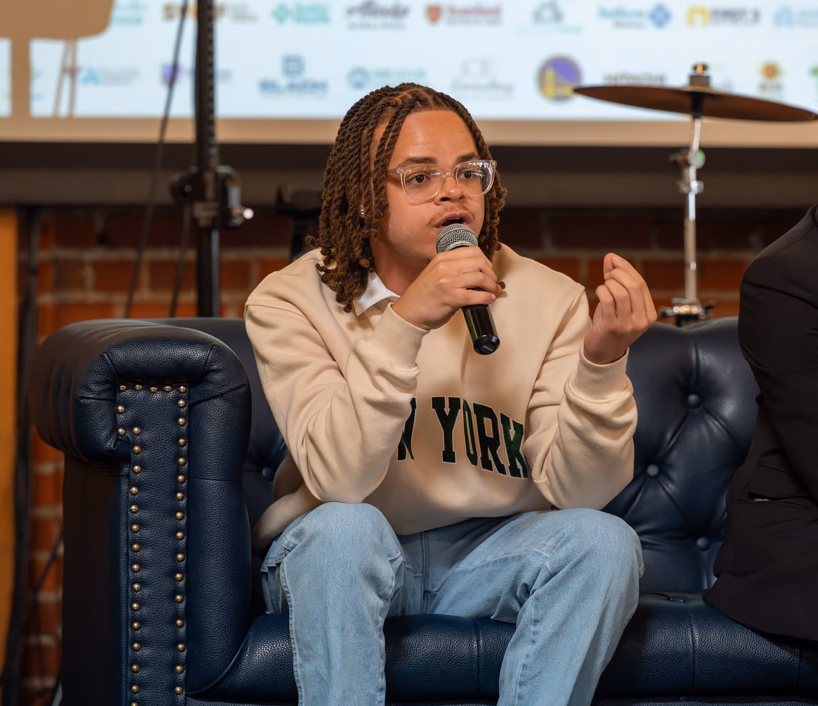 A panelist wearing clear glasses and a New York sweatshirt speaks into a microphone from a blue couch