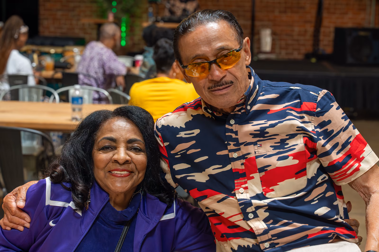 Two guests pose closely together indoors, one in a purple jacket and the other wearing amber glasses