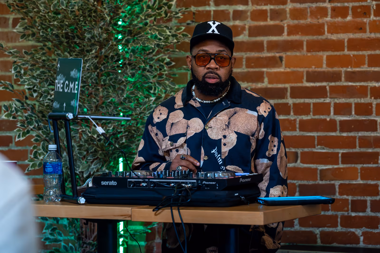 A DJ in a patterned shirt and black cap stands behind a mixing console against a brick wall