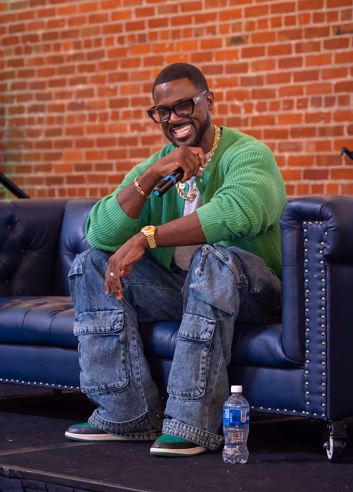 A smiling speaker in a green cardigan leans forward with a microphone on a couch against a brick wall