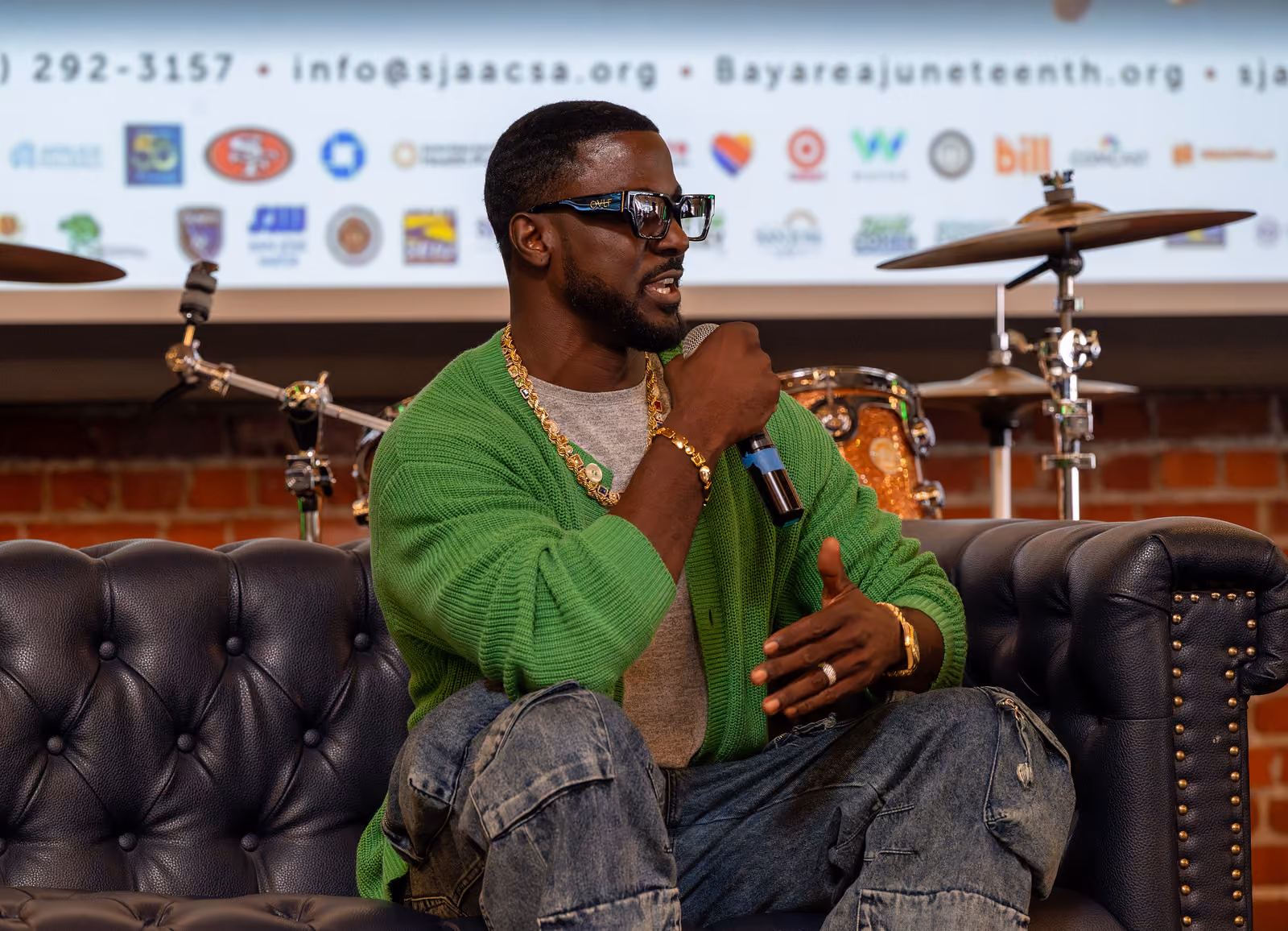 A speaker in a green cardigan holds a microphone while seated on a dark tufted couch near a drum kit