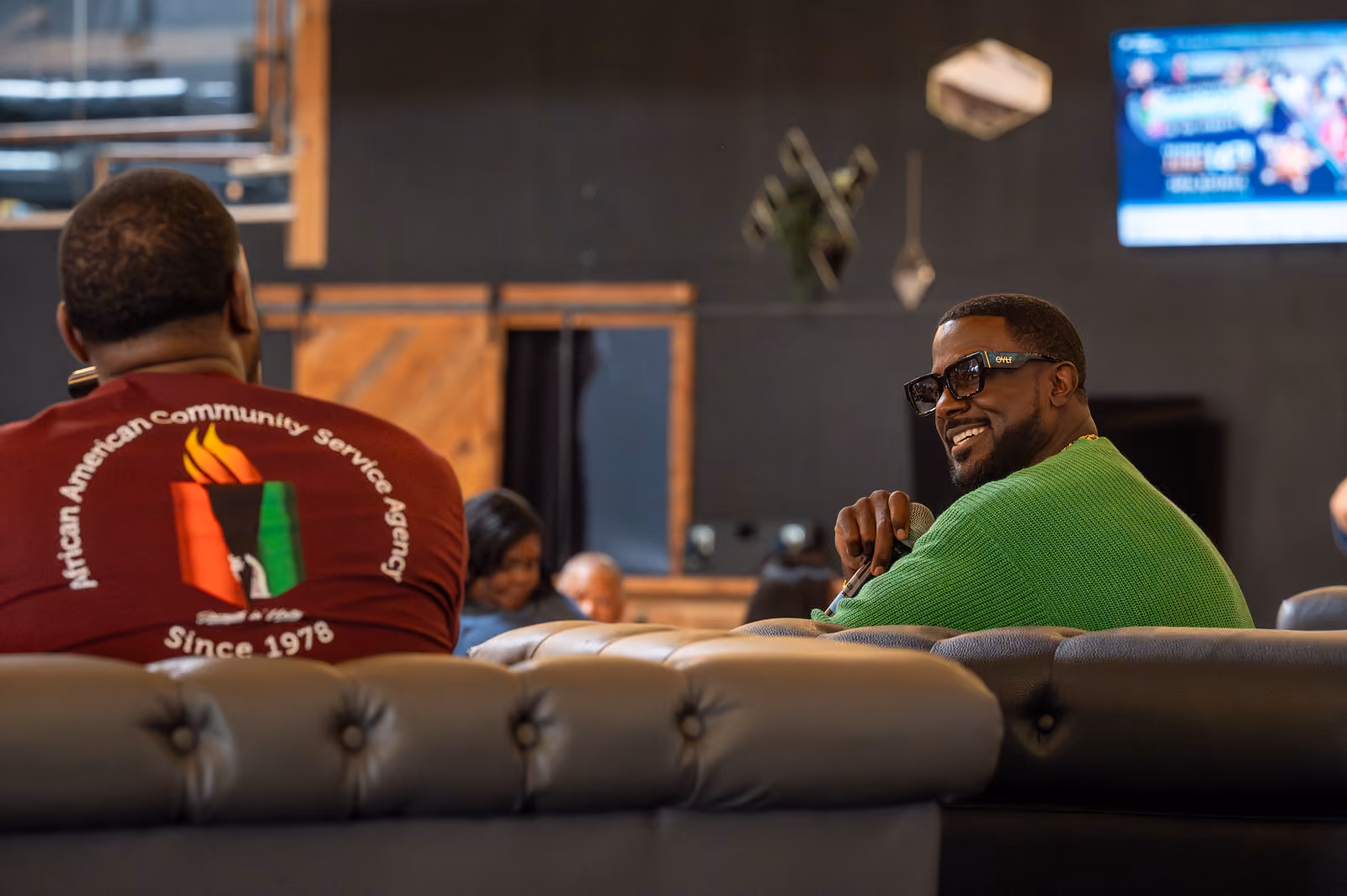 A seated speaker in a green cardigan smiles toward a guest wearing an AACSA shirt across the couch