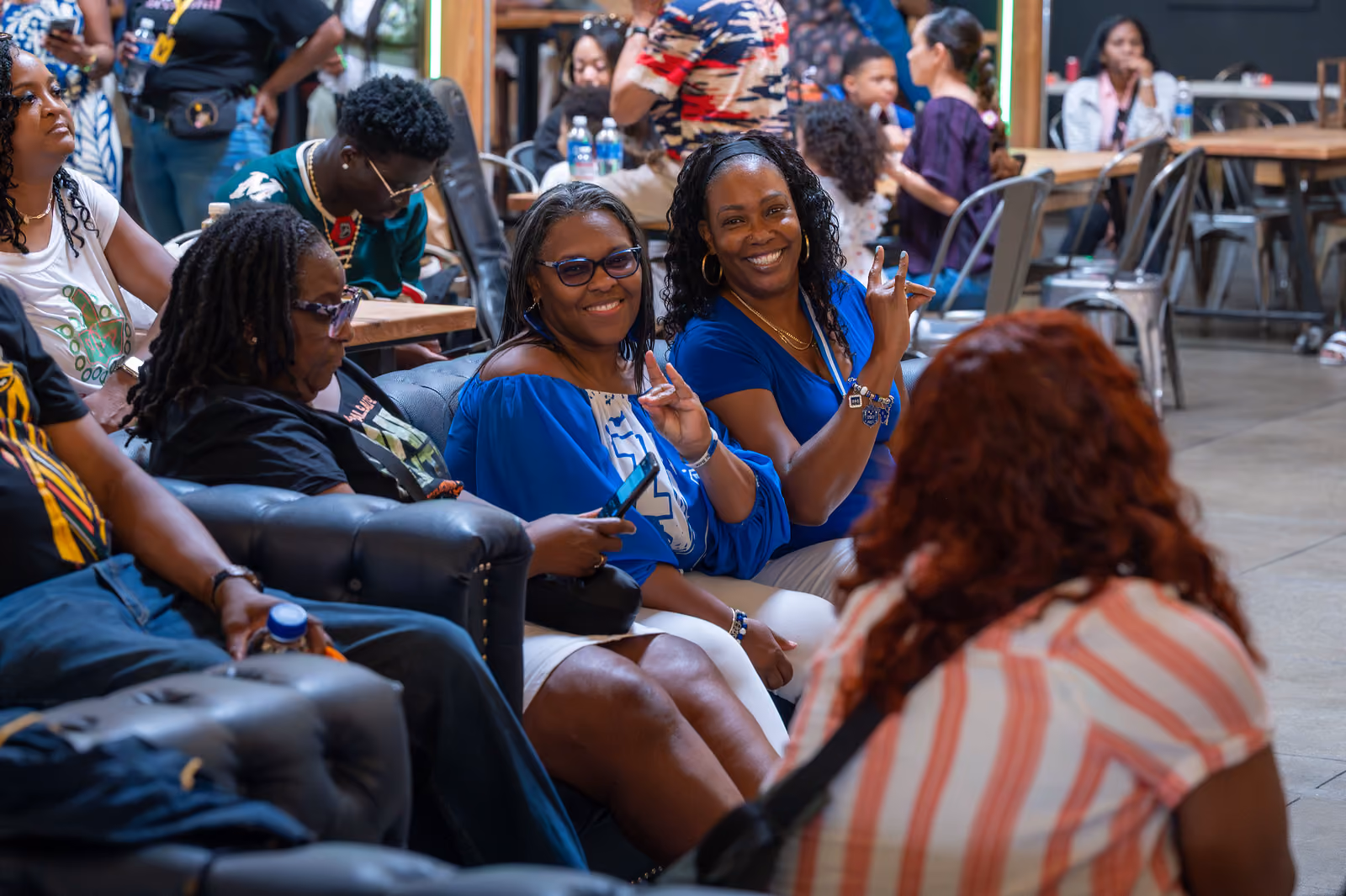 Two smiling guests in blue tops wave toward the camera while seated among festival attendees indoors