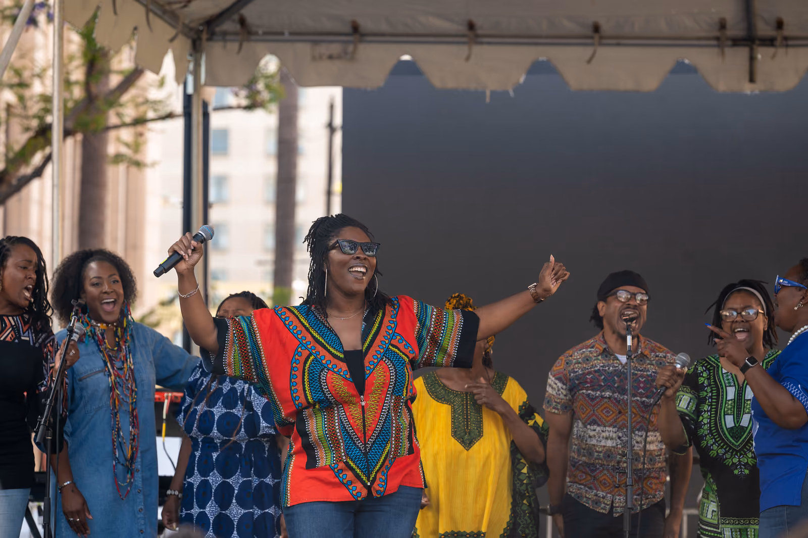 A singer in a red patterned shirt spreads her arms with a microphone as fellow singers perform onstage
